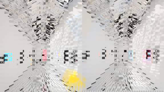 A bright, white-walled art gallery with a striped floor, several small paintings, a central yellow sculpture, and a sloped, skylight ceiling.