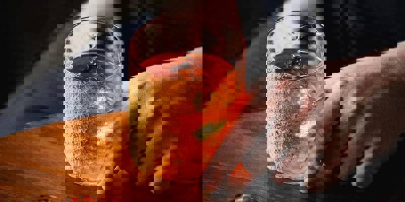 A hand holds an old-fashioned cocktail with a crumb-coated rim next to an orange-glowing tea light on a wooden table.