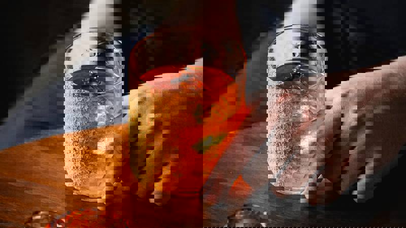 A hand holds an old-fashioned cocktail with a crumb-coated rim next to an orange-glowing tea light on a wooden table.