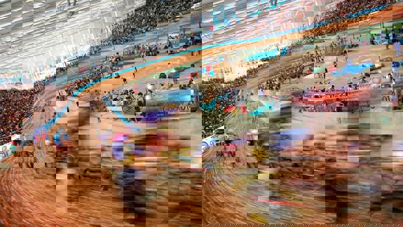 A wide-angle, blurred shot of a cycling race inside a velodrome. Cyclists are racing around a large, wooden track. The stands are filled with spectators.