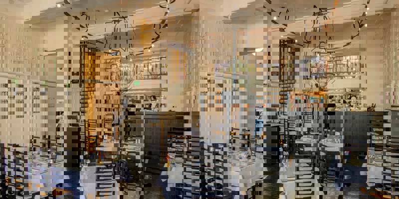 The interior of a Mackintosh-designed tearoom, featuring distinctive high-backed chairs, white tablecloths, and unique lighting fixtures, with a staircase in the background.