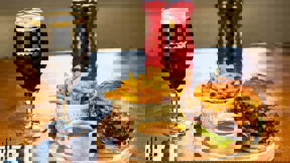 A hearty burger with melted cheese and lettuce, a bowl of chips, gravy, and a dark beer in a stemmed glass on a wooden table.