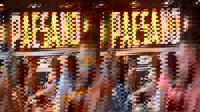 The interior of a busy Italian restaurant, with a large, lit-up 'PAESANO' sign on the wall behind people dining at tables.