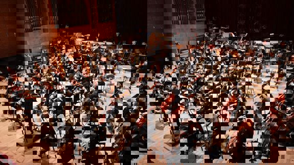 Large orchestra of students from the Royal Conservatoire of Scotland performing on stage, with conductor leading string and brass sections.