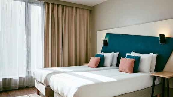 A bright hotel room with two single beds dressed in white, featuring a teal headboard and accent pillows. Sunlight streams through sheer window curtains.