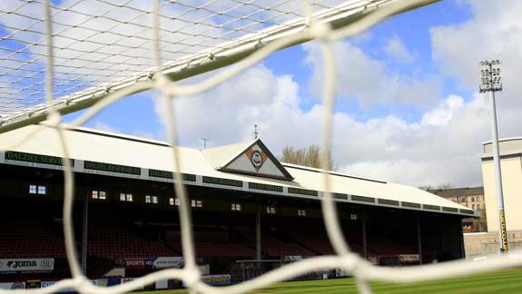 The stadium stands of a football ground are seen through the white netting of a goal. The main stand has a white roof with a triangular logo.