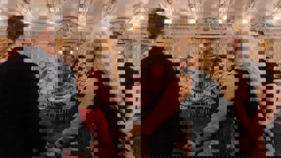 People dance in a circle at a ceilidh, a traditional Gaelic social gathering, in a room with ornate ceilings and chandeliers.