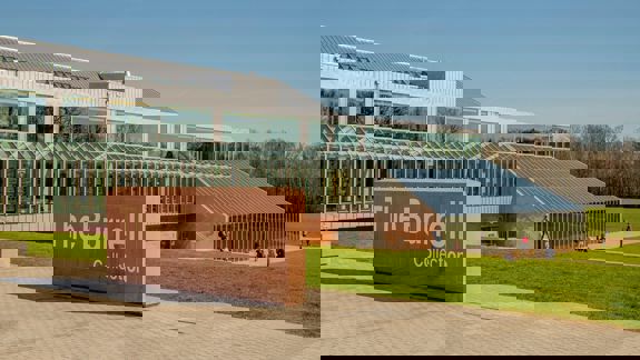 The modern, glassy Burrell Collection building is shown on a sunny day with people walking on the surrounding lawn. A large stone sign with the name of the building is in the foreground.