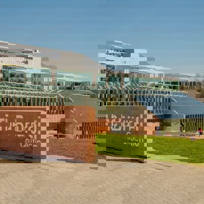 The modern, glassy Burrell Collection building is shown on a sunny day with people walking on the surrounding lawn. A large stone sign with the name of the building is in the foreground.
