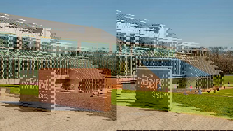 The modern, glassy Burrell Collection building is shown on a sunny day with people walking on the surrounding lawn. A large stone sign with the name of the building is in the foreground.