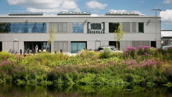 A two-story, modern building with a corrugated metal exterior sits next to a body of water, surrounded by tall grasses and purple flowers.