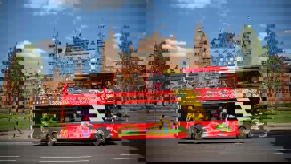 A red open-top sightseeing bus parked in front of the Kelvingrove Art Gallery and Museum in Glasgow on a sunny day.