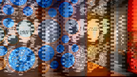 An interior shot of an exhibition with large circular art pieces on a wall, some with text like "CITY GLASGOW 1903".