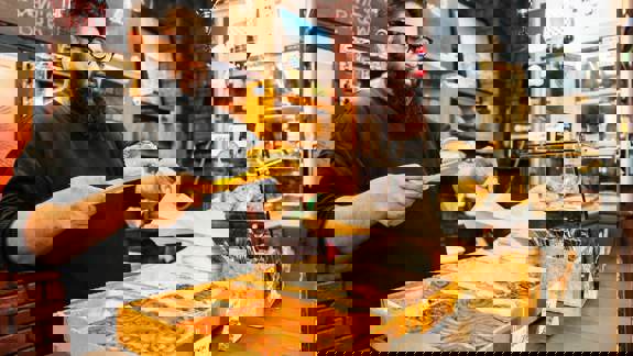 A person with a beard and glasses uses yellow tongs to place a pastry into a paper bag. They stand behind a counter  displaying trays of cinnamon rolls and other baked goods inside Ottoman Coffee House. 