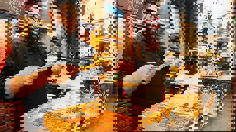 A person with a beard and glasses uses yellow tongs to place a pastry into a paper bag. They stand behind a counter displaying trays of cinnamon rolls and other baked goods inside Ottoman Coffee House.