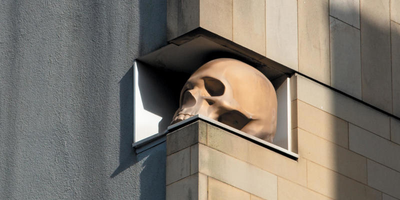 A realistic, oversized human skull sculpture rests within a rectangular cut-out of a modern building.