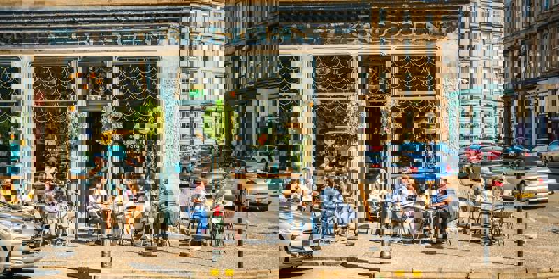 People sit at outdoor tables on a sunny day in front of a cafe with large windows and hanging plants.