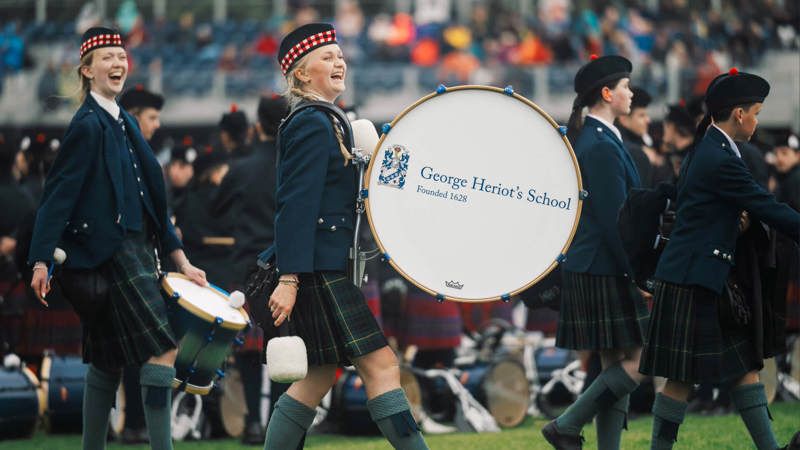 Smiling members of a marching band in traditional Scottish attire perform with drums and bagpipes at an outdoor event, with crowds in stadium seating visible in background.
