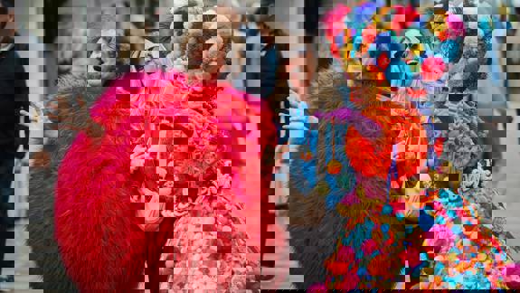 Two street performers in vibrant, colourful costumes, one in a red fuzzy suit and another in a multi-coloured pom-pom dress, pose with a woman.