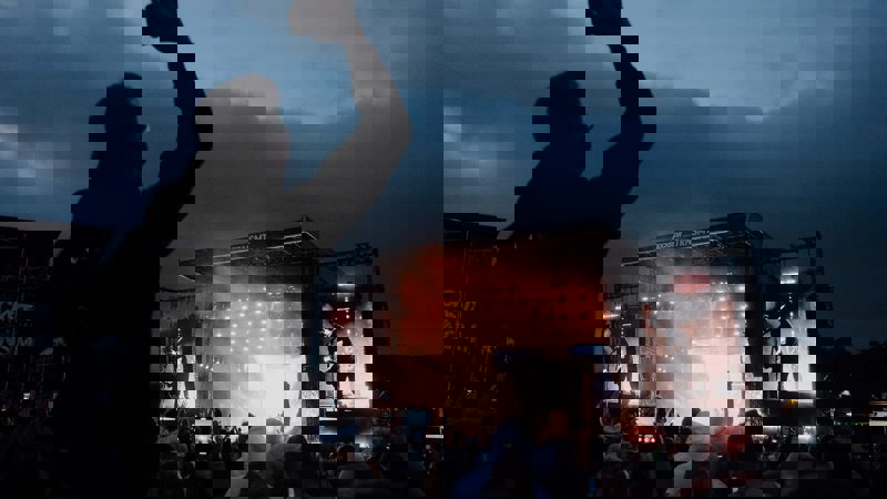 A dark shot of a crowd from behind watching a stage at a music festival. One person in the crowd stands out, as they are sitting on another person’s shoulders.