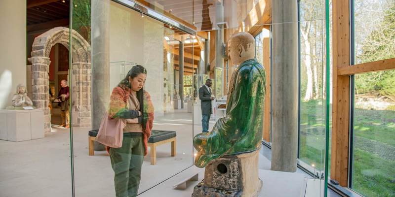 A person looks at a large seated statue in a glass case inside a modern museum with tall windows and wooden beams.