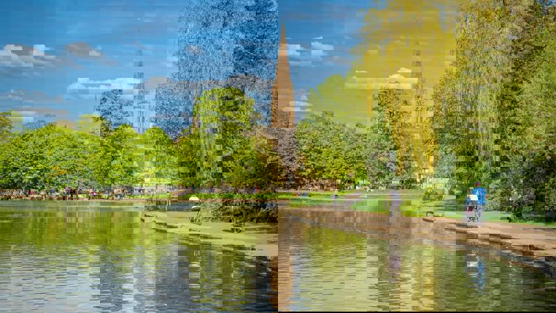 A large pond in a park with a tall church steeple in the background, with people walking and cycling on a path.