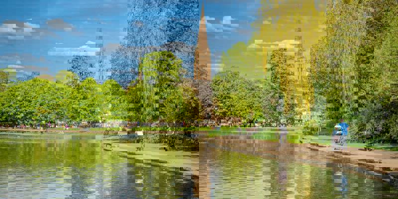 A large pond in a park with a tall church steeple in the background, with people walking and cycling on a path.