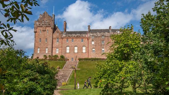 The grand red sandstone facade of Brodick Castle, featuring historic battlements and turrets, set behind lush green trees and a sloping lawn with a stone staircase.