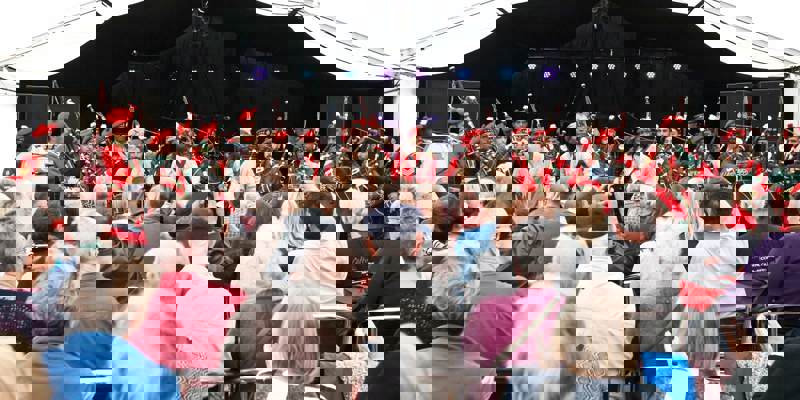 An audience watches a performance by a pipe band, dressed in red and green uniforms and turbans, on a stage under a large white tent.
