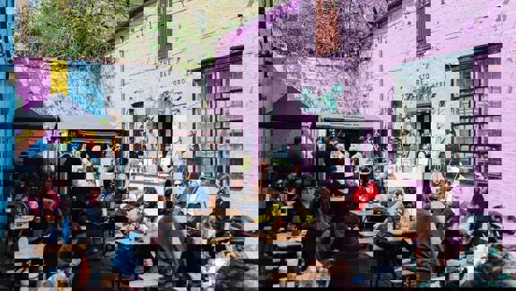 A bustling outdoor market scene with many people sitting at picnic tables in a courtyard with bright pink and yellow walls.