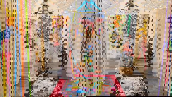  An art installation with dolls, colourful textile hangings, and figures, including one on a red rug. A figure in a white dress stands beside it.