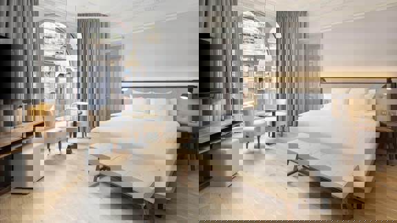 Stylish guest room at AC Hotel Glasgow with a large bed, modern lighting, light wooden floors, and a seating area beside tall arched windows overlooking the city.