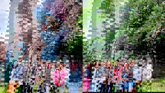 A group of people standing in front of the iconic "St Mungo" mural by Smug on High Street, Glasgow. The large-scale artwork depicts a modern-day man in a beanie with a robin.