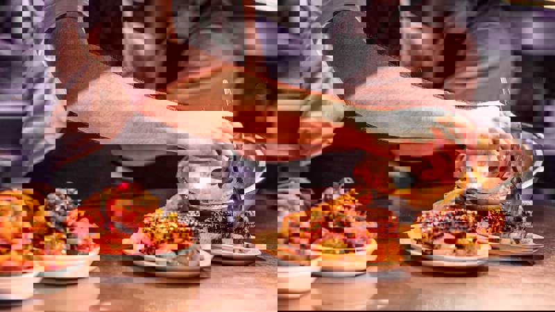 A chef's hands are plating dishes of roasted corn on the cob and other food on a metal counter in a restaurant kitchen.