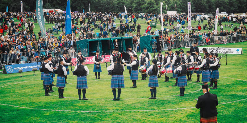  pipe band in kilts performs in a grassy field, surrounded by a large crowd of spectators at the World Pipe Band Championships.