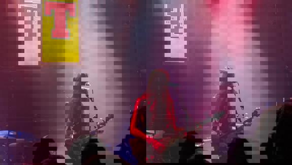 A female musician with long dark hair plays a black electric guitar and sings into a microphone on a stage bathed in red and white spotlights. A yellow and red 'T' banner is visible.