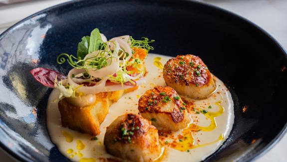 Close-up of three seared scallops and a potato cube in a creamy sauce in a dark blue bowl.