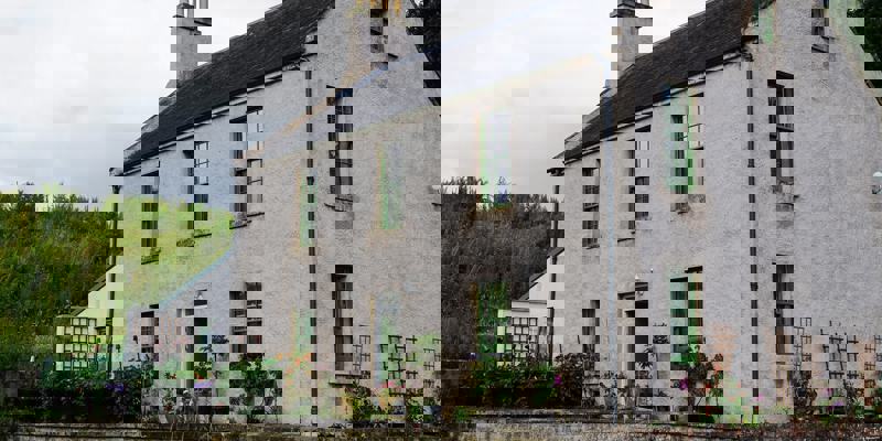 A two-story, grey stone house with green-framed windows, surrounded by a low brick wall and lush green plants. In the foreground, there are three colorful chairs and a small chalkboard.