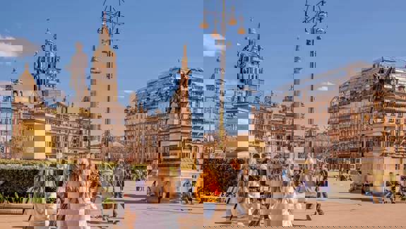George Square, Glasgow, on a sunny day with people walking and statues prominent, including the City Chambers building in the background.