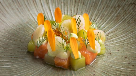 A close-up of a meticulously plated, modern dish of salmon and green cubes, topped with orange petals and fish roe.