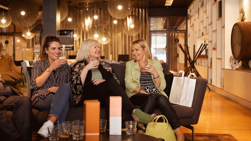 Three people sit on a sofa, smiling and holding drinks. They are sitting in a well-lit room with hanging lights and bottles on the table.