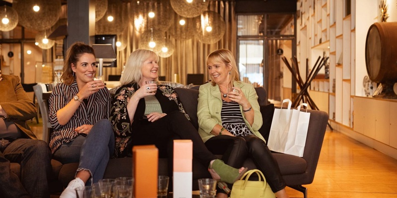 Three people sit on a sofa, smiling and holding drinks. They are sitting in a well-lit room with hanging lights and bottles on the table.