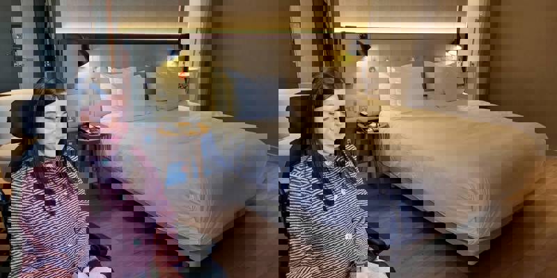 A person in a wheelchair sitting in a modern hotel room, holding a cup and looking down, with a large bed in the background.