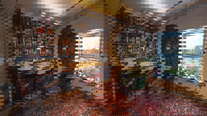 The studio of Alasdair Gray, showing a room with a detailed desk, bookshelves filled with books, a large green armchair, and a window overlooking the city.