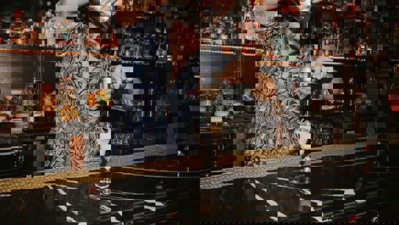 A bartender in a patterned shirt strains a cocktail into a glass on a black marble bar, with shelves of bottles behind him.