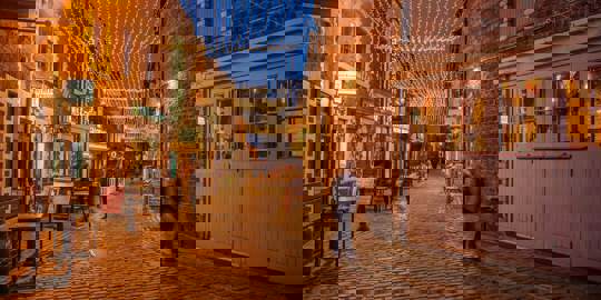 A cobblestone lane in Glasgow, Ashton Lane, lit by hanging fairy lights and lanterns, with people walking along it at dusk.