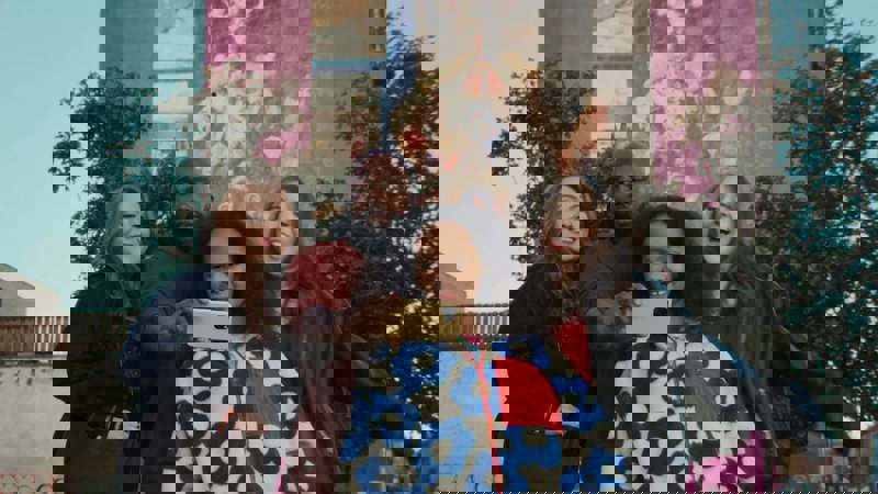 A group of four smiling friends take a selfie together outdoors. They stand before a pink building featuring a large mural, surrounded by green trees under a clear sky.